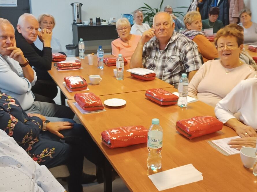 Grupa seniorów na szkoleniu z pierwszej pomocy, siedzą przy stołach z apteczkami i wodą.