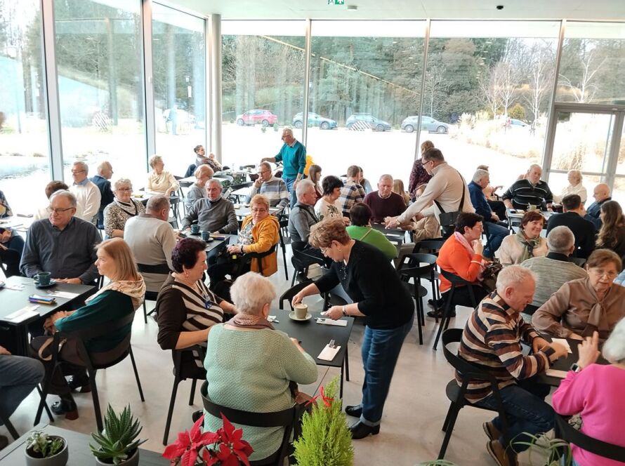 Wnętrze kawiarni z dużą grupą ludzi rozmawiających przy stolikach.