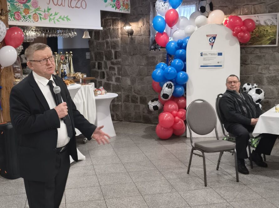 Mężczyzna w garniturze przemawia na przyjęciu z balonami i dekoracjami.