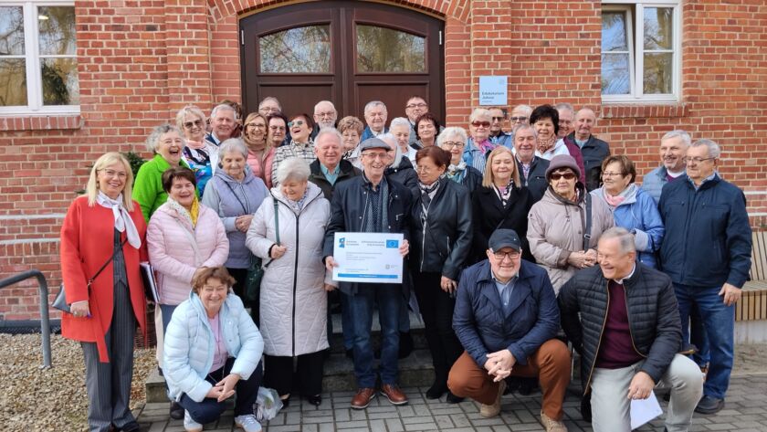 Grupa starszych osób przed budynkiem z cegły, trzymających certyfikat.