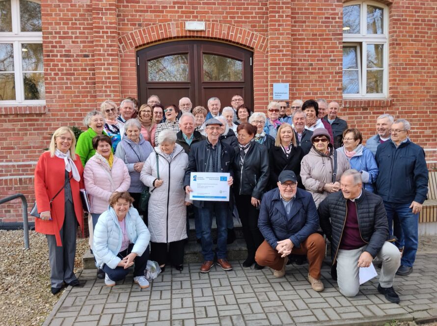 Grupa starszych osób przed budynkiem z cegły, trzymających certyfikat.