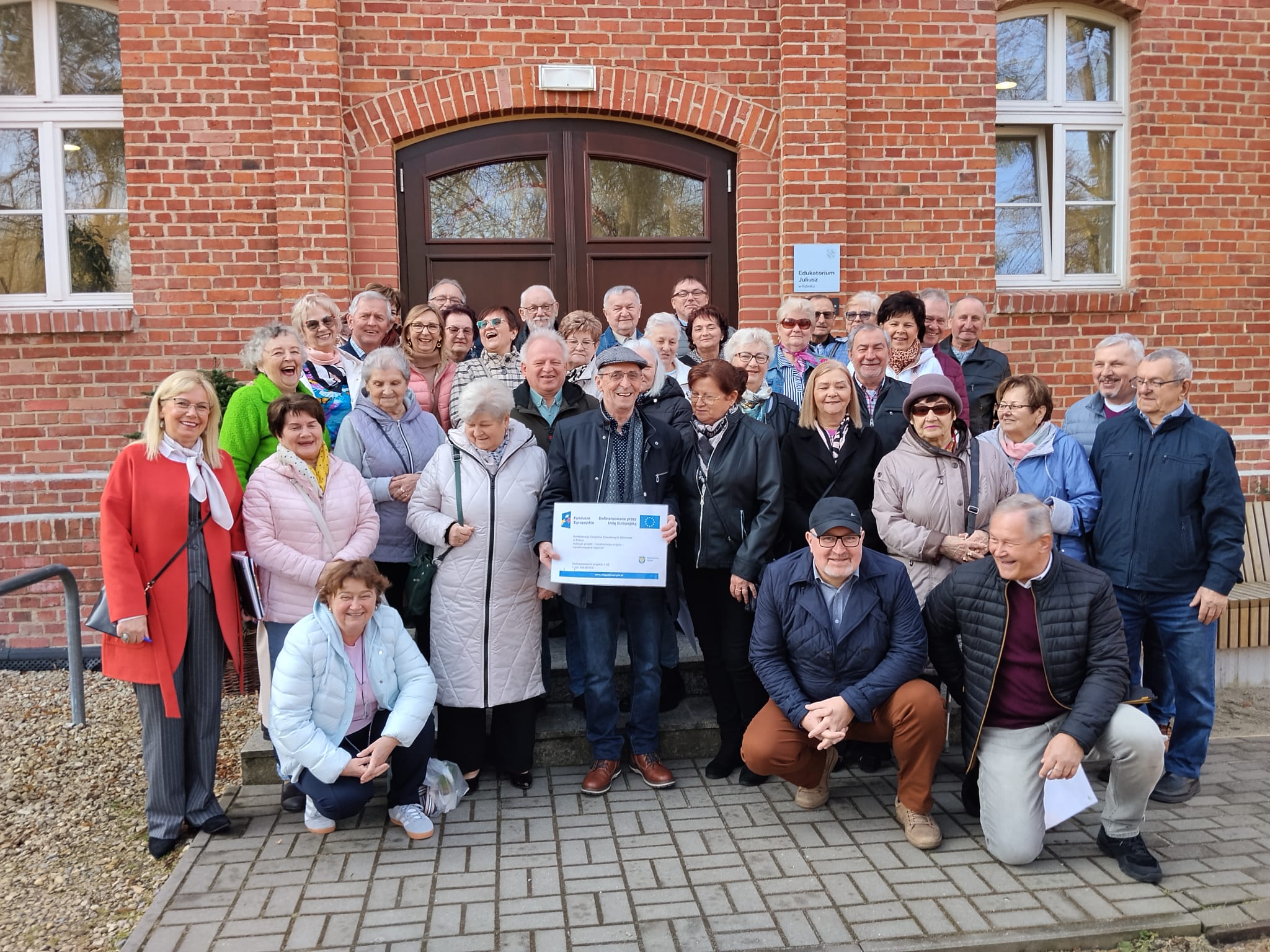 Grupa starszych osób przed budynkiem z cegły, trzymających certyfikat.