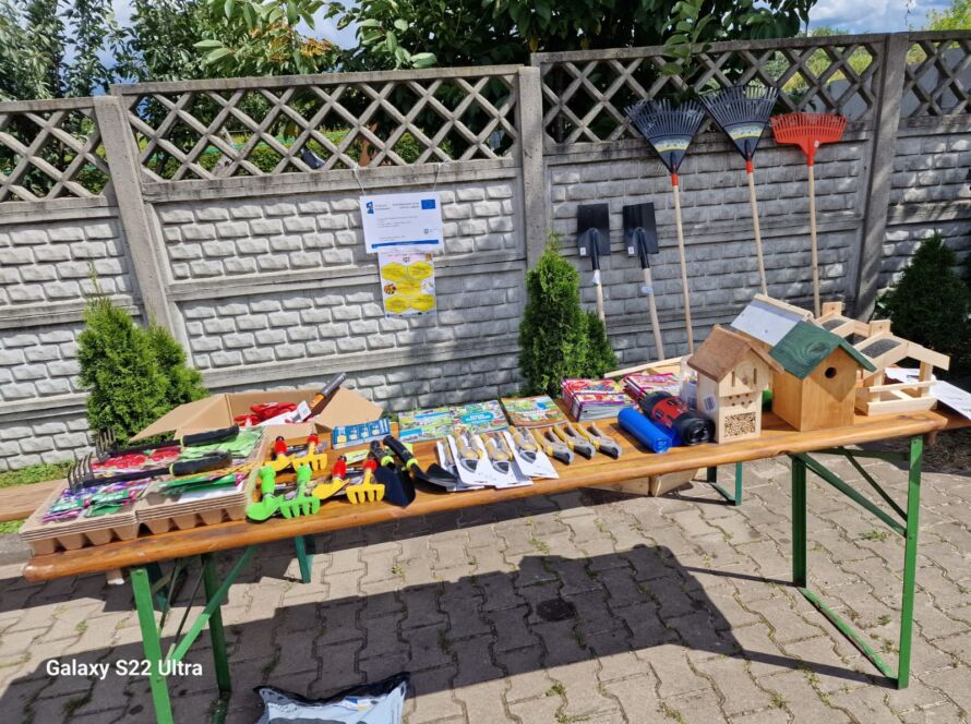 Stół z narzędziami ogrodniczymi, karmnikami i broszurami na tle ogrodzenia.