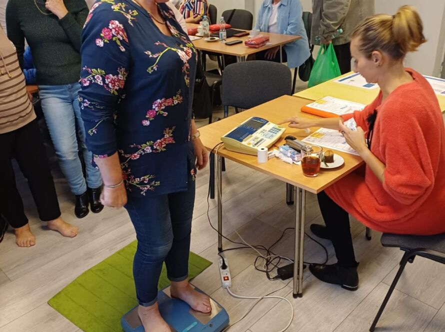 Kobieta stoi boso na wadze podczas badania w sali, obok osoba przy stole notuje wyniki