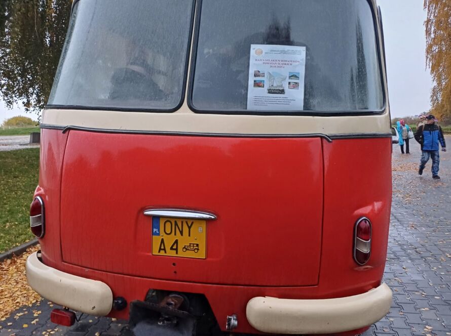 Tył klasycznego czerwono-kremowego autobusu na deszczu, ulotka na szybie, jesienne liście na bruku.