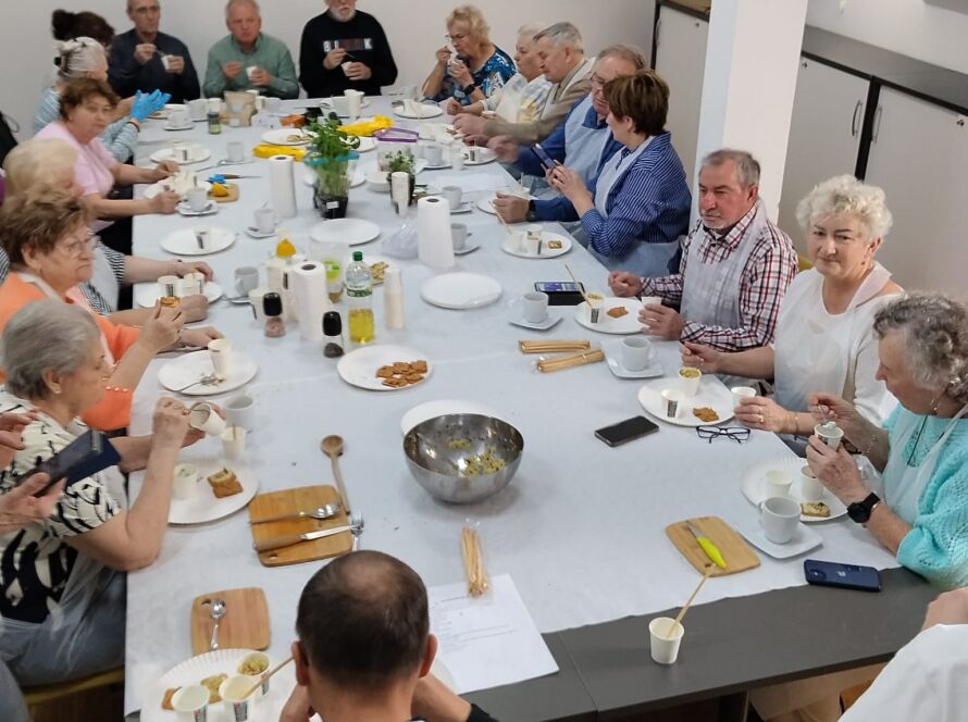Grupa osób siedzących przy wspólnym stole, jedzących i rozmawiających.
