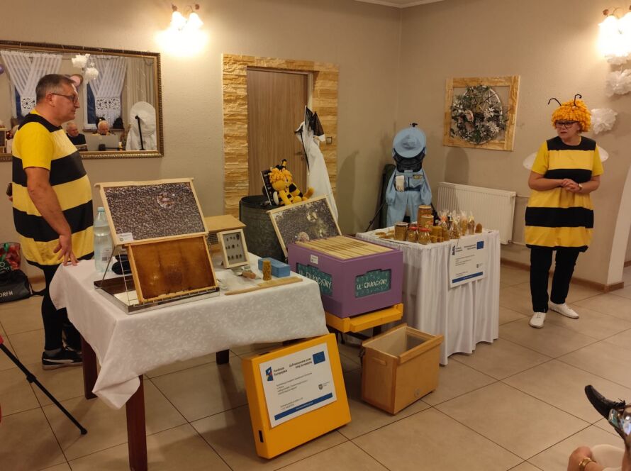 Osoby w kostiumach pszczół na stoisku edukacyjnym z produktami pszczelarskimi.