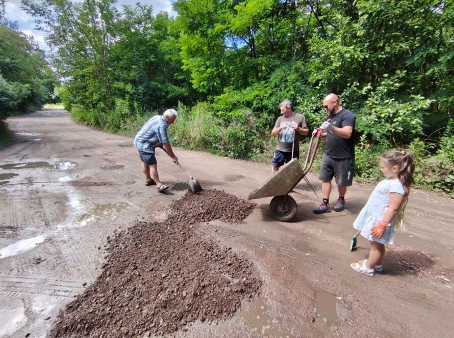 Grupa osób wypełniająca dziury na drodze ziemią w leśnym otoczeniu.
