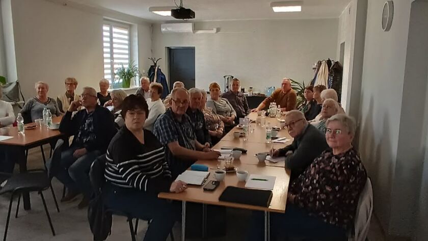 Grupa starszych osób siedzących przy stołach podczas spotkania w sali.