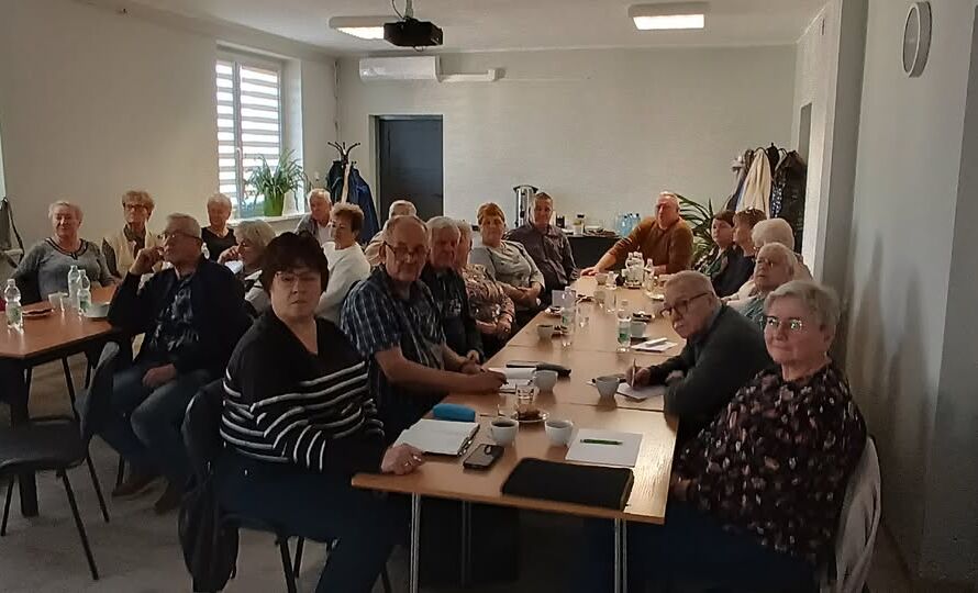 Grupa starszych osób siedzących przy stołach podczas spotkania w sali.