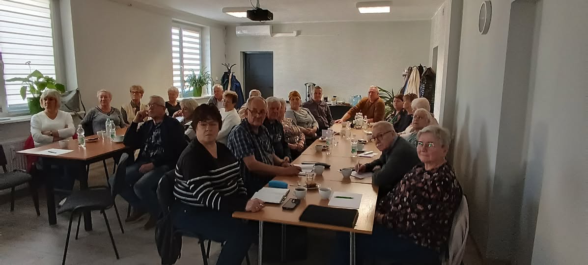 Grupa starszych osób siedzących przy stołach podczas spotkania w sali.