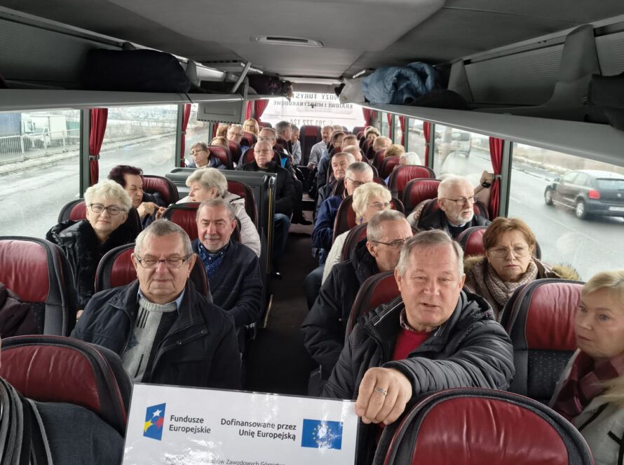 Grupa starszych ludzi siedzi w autobusie trzymając znak Funduszy Europejskich.