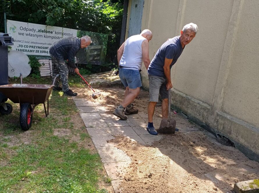 Mężczyźni pracujący na podwórku, przesiewają piasek w pobliżu kompostownika.