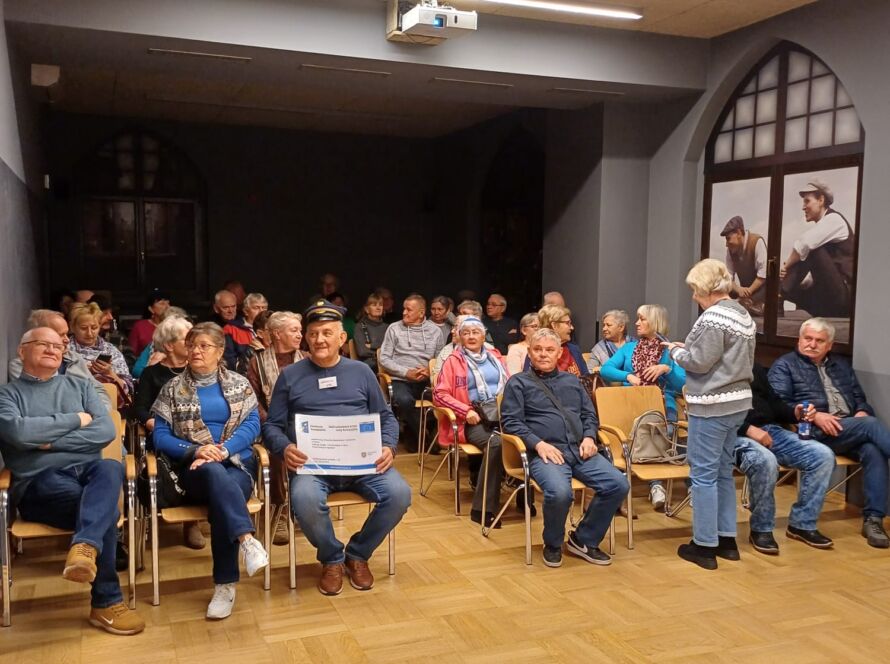 Grupa dorosłych siedzi w sali z projektorem; część rozmawia, a jeden mężczyzna trzyma plakat, oczekując na spotkanie.