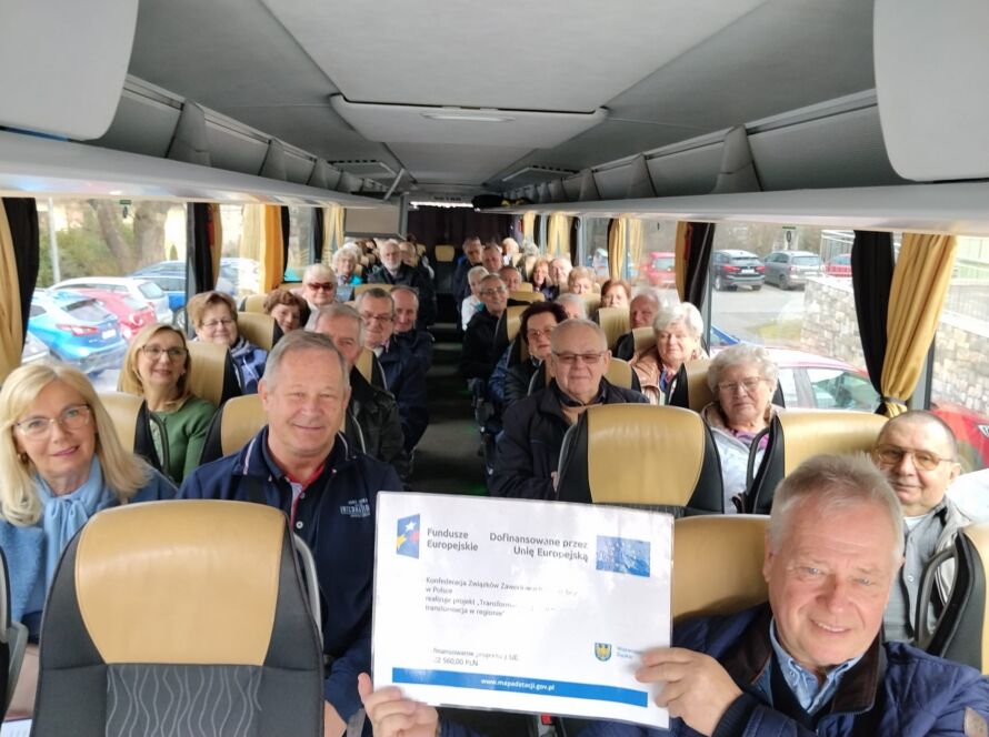 Seniorzy siedzący w autobusie, uśmiechnięci, trzymający plakat.