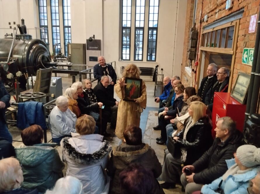 Grupa osób w muzeum, przewodnik z obrazem mówi do zwiedzających.