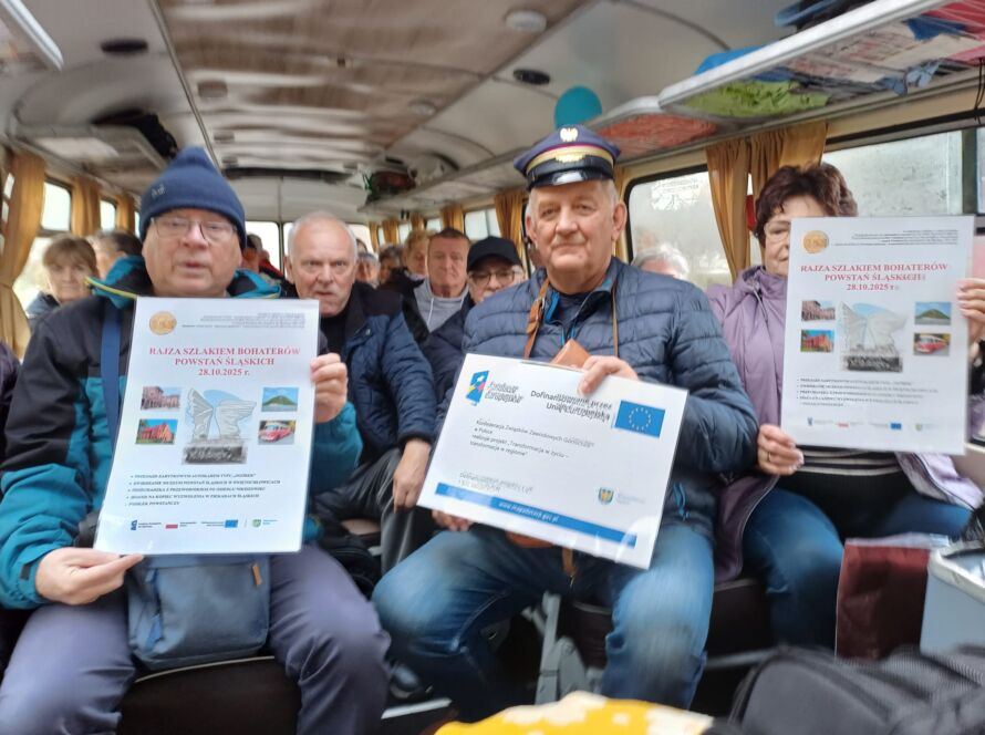 Grupa dorosłych w autobusie trzyma plakaty Rajzy Szlakiem Bohaterów Powstań Śląskich.
