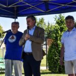 Trzech mężczyzn stoi pod niebieskim namiotem na świeżym powietrzu, dwóch mężczyzn trzyma mikrofony.