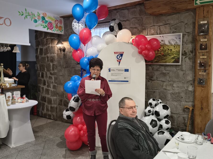 Kobieta przemawiająca z mikrofonem obok dekoracji z balonów i piłek.