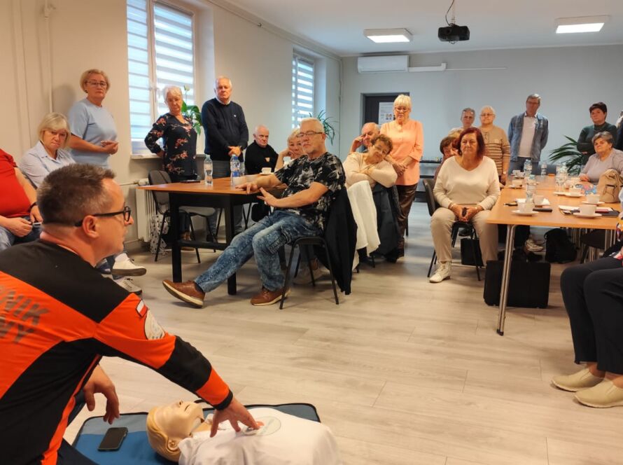 Ratownik medyczny pokazuje RKO na fantomie podczas szkolenia, a grupa seniorów uważnie obserwuje w sali.