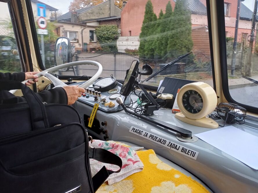 Wnętrze kabiny kierowcy zabytkowego autobusu z retro zegarami, żółtym kocem i widokiem na deszczową ulicę.