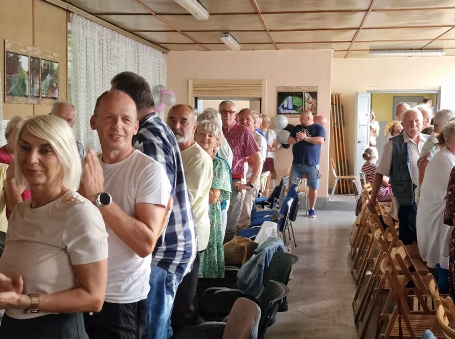 Ludzie ustawieni w kolejce w sali z krzesłami i dekoracjami.