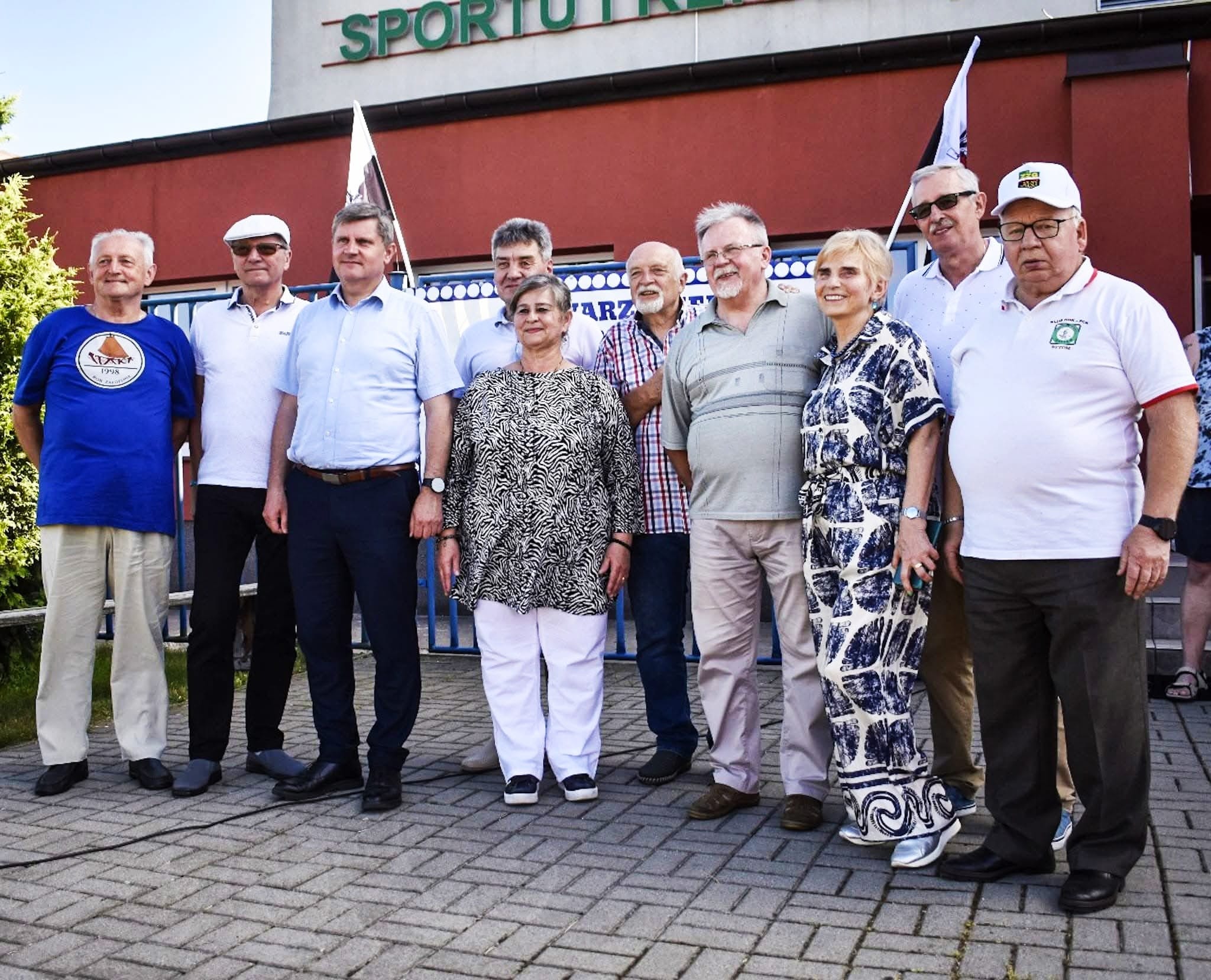 Grupa starszych osób stojąca przed budynkiem sportowym.