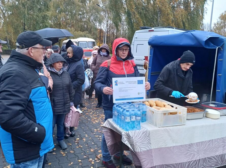 Ludzie stoją w kolejce przy mobilnym stoisku z jedzeniem; na stole bułki i woda, jesienny deszcz.
