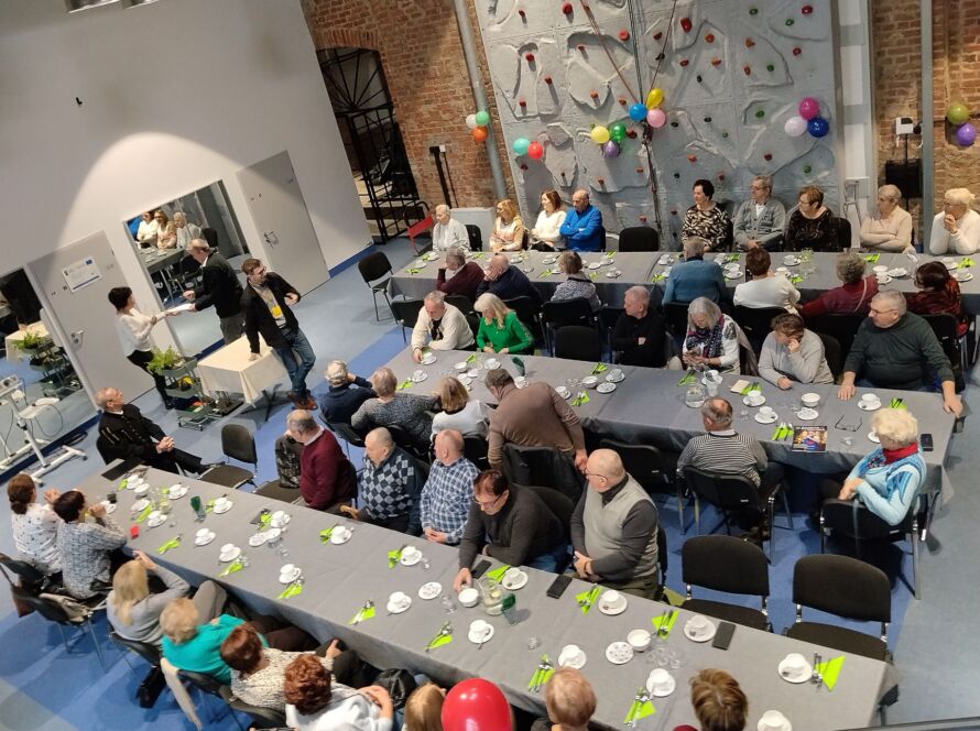 Spotkanie osób przy długim stole z kawą i ciastem, balony i ściana wspinaczkowa w tle.