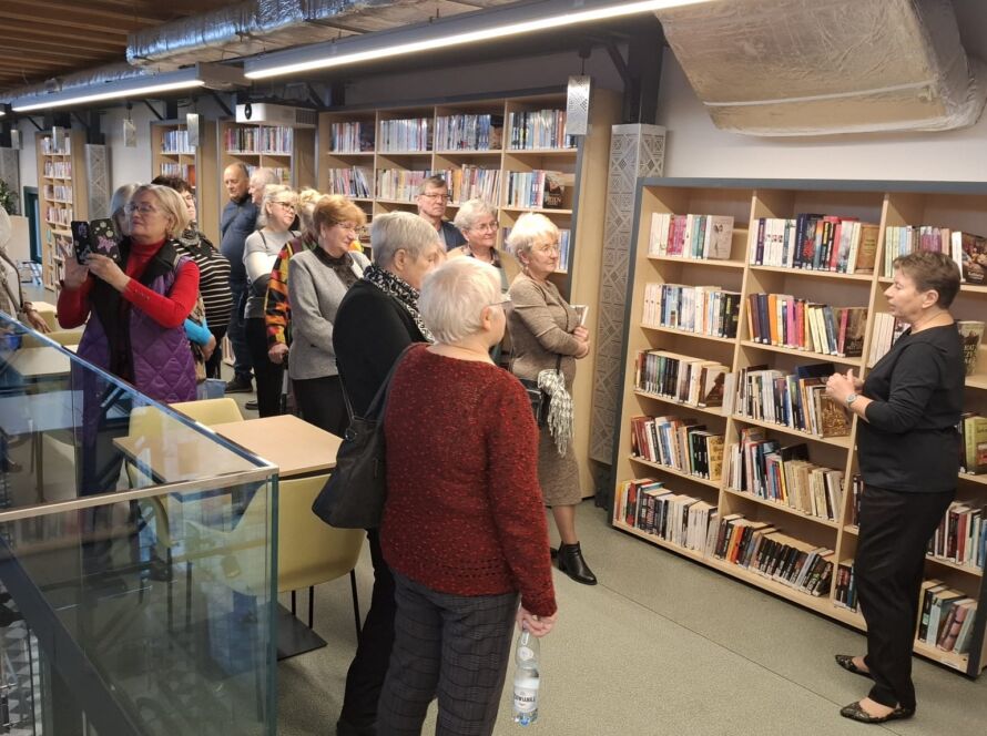 Grupa ludzi zwiedza bibliotekę, przewodniczka przy półce z książkami.