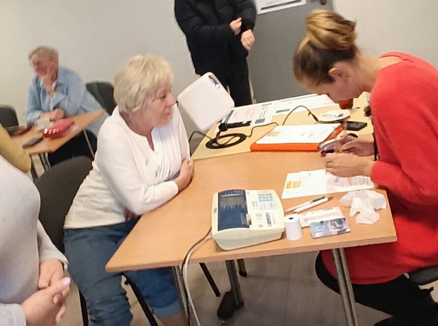 Konsultacja zdrowotna w sali: kobieta zapisuje wyniki, seniorzy czekają, ciśnieniomierz na stole