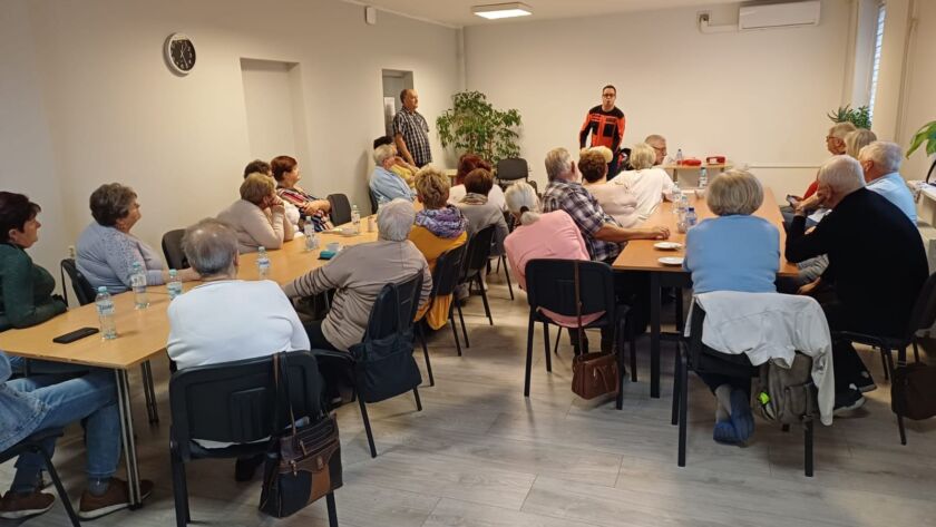 Grupa seniorów na spotkaniu w sali; prowadzący stoi z przodu, woda na stołach.