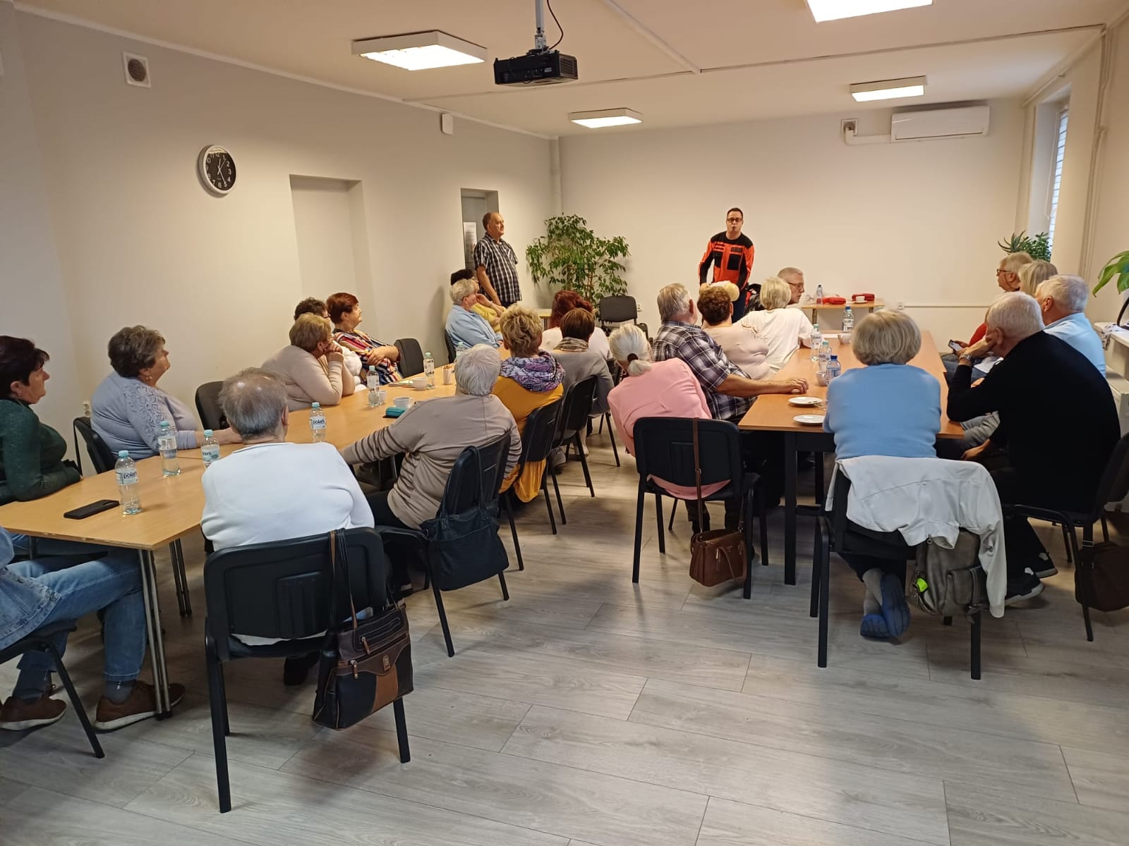 Grupa seniorów na spotkaniu w sali; prowadzący stoi z przodu, woda na stołach.