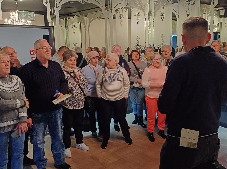 Grupa seniorów słucha przewodnika w eleganckiej sali z kryształowym żyrandolem i lustrami.