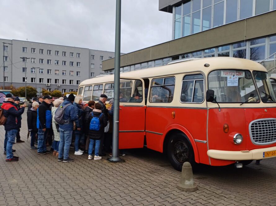 Grupa ludzi w kolejce do zabytkowego czerwono-kremowego autobusu przed nowoczesnym budynkiem.