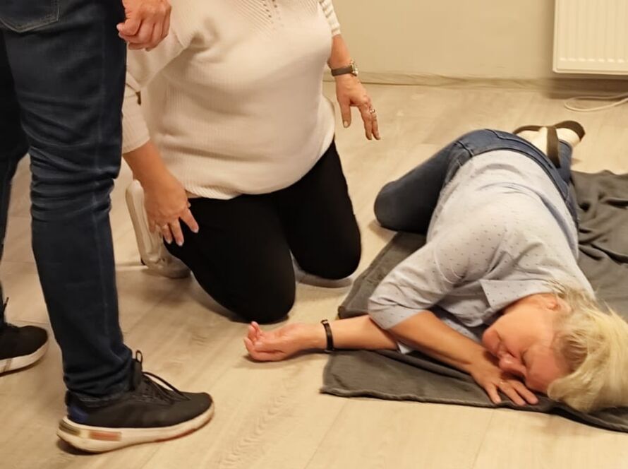 Szkolenie z pierwszej pomocy: kobieta w pozycji bocznej na kocu, dwie osoby pomagają.